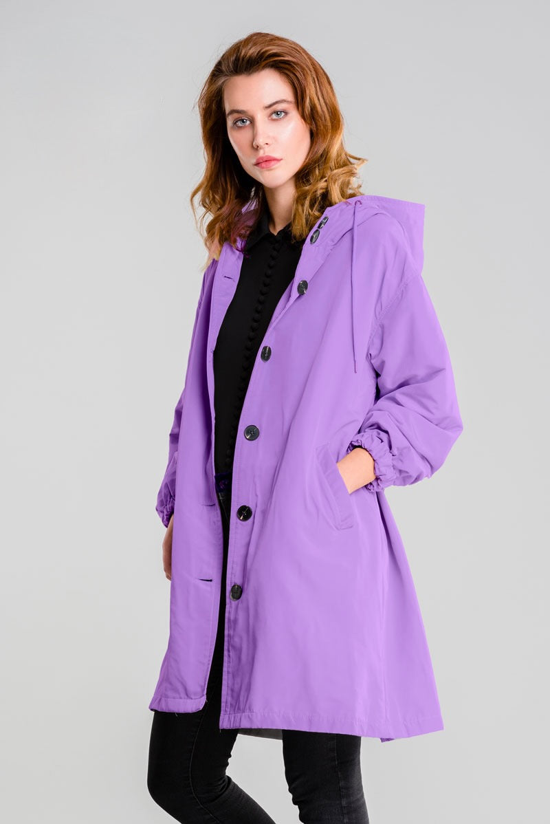 A person wears the looselyboho SOFT LAVENDER WINDBREAKER featuring black buttons, pockets, and an adjustable hood, standing against a plain light gray background.