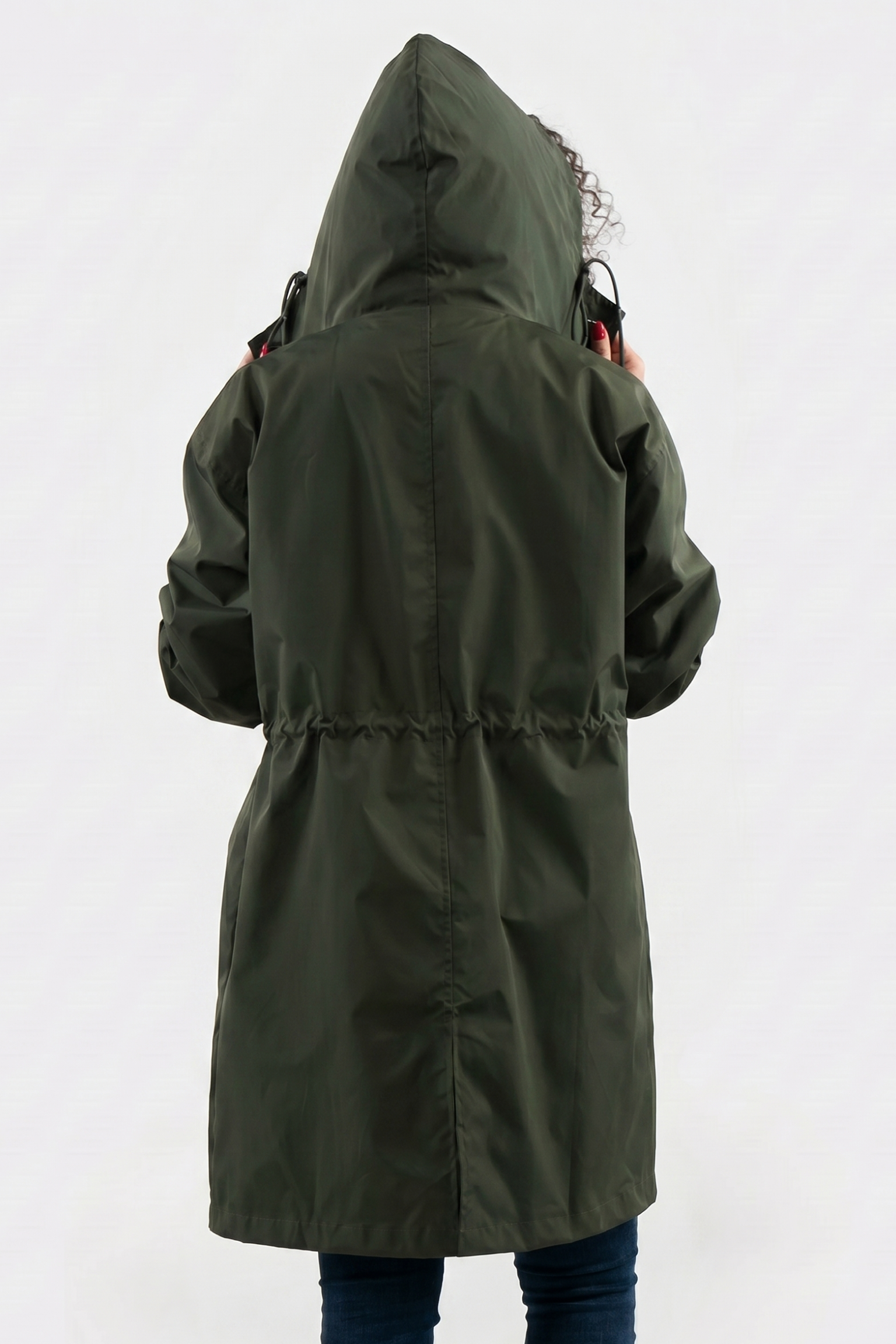 A person in blue jeans is shown from the back wearing the Icon Windbreaker Rain Jacket in olive green with the hood up, adjusting it. The background is plain and light-colored.