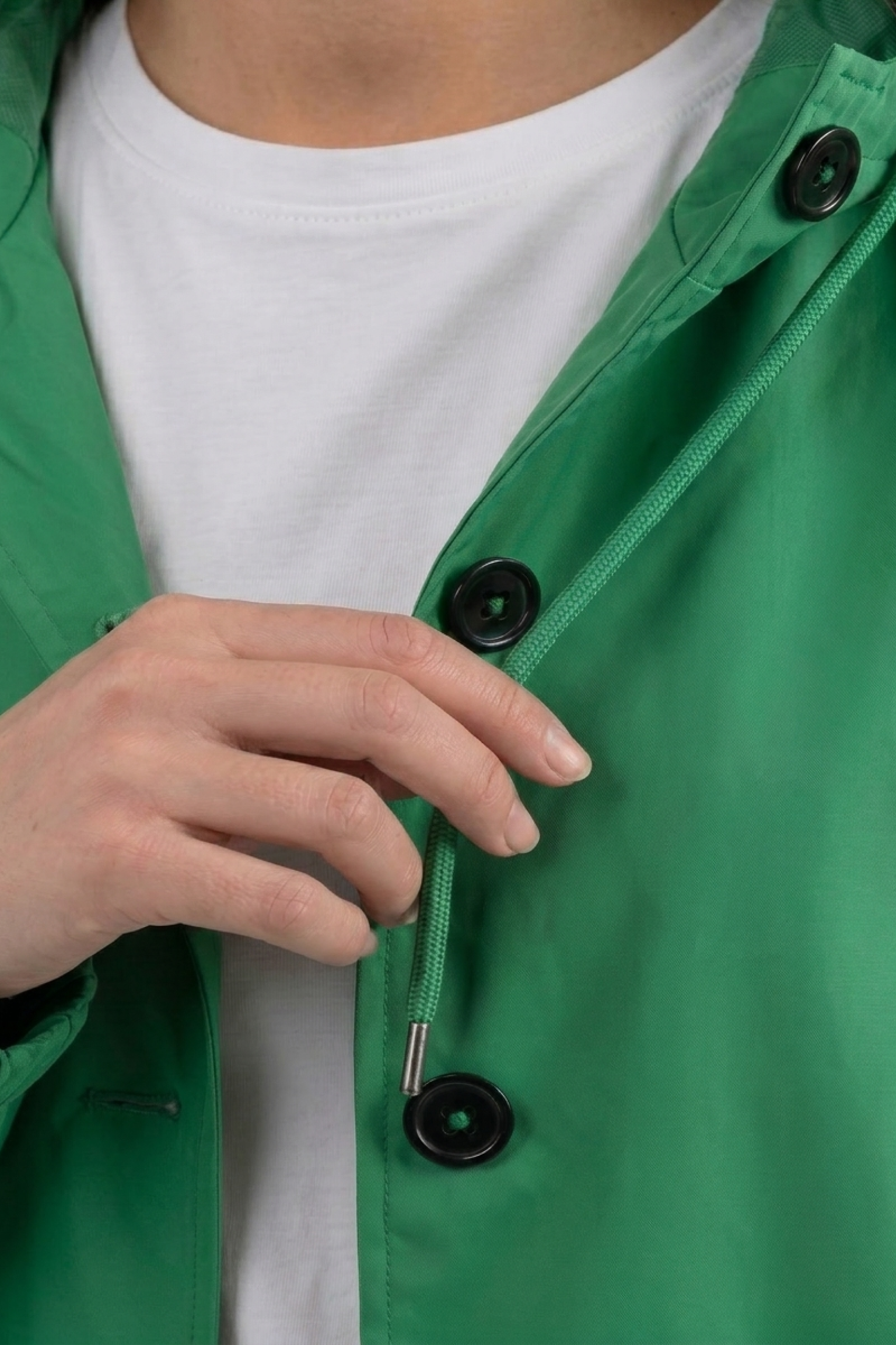 A person wearing a white shirt and the Icon Windbreaker Rain Jacket in green fastens a black button on the jacket with their left hand.