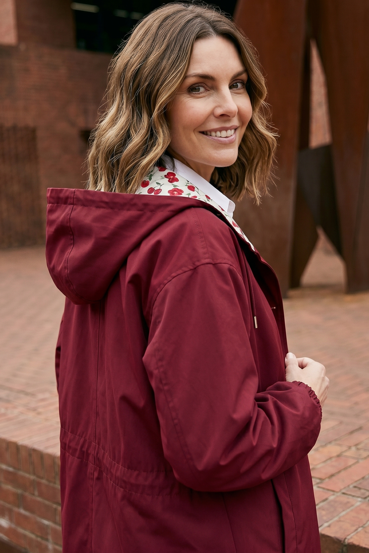 A woman with wavy brown hair wears the Icon Windbreaker Rain Jacket and smiles at the camera while standing outdoors on a brick walkway, with brick walls and metal structures in the background.