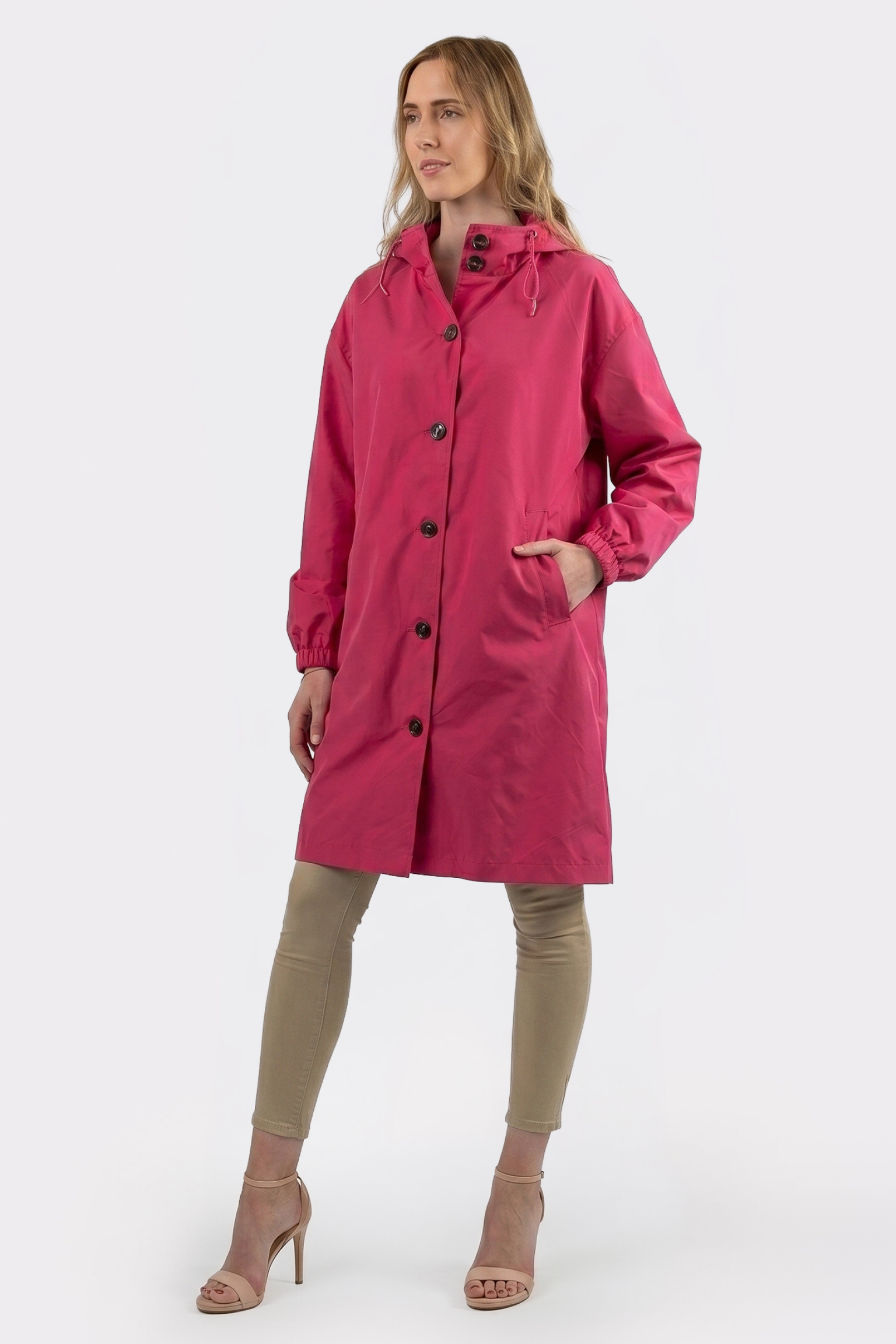 A person is wearing the Icon Windbreaker Rain Jacket—a long pink button-up coat with a hood—paired with beige leggings and beige high-heeled sandals, standing against a plain light background.