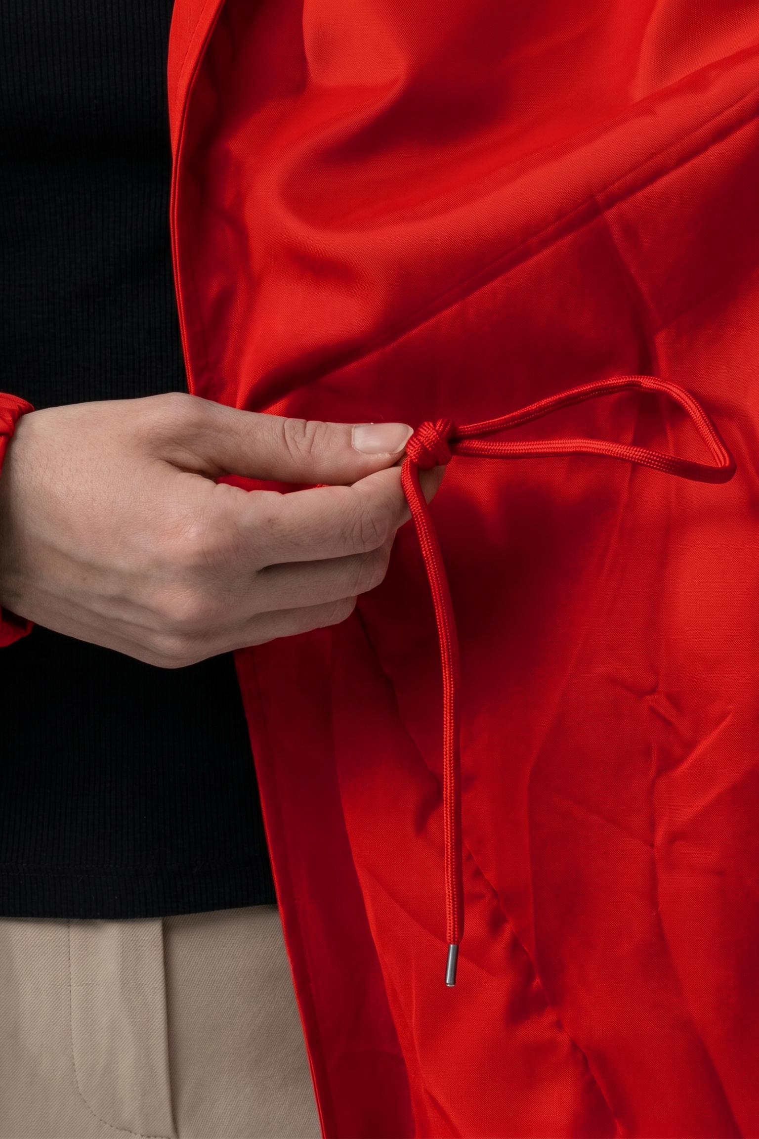 A person tightens the red drawstring of the Icon Windbreaker Rain Jacket while wearing a black shirt and beige pants. Only their hand, part of their torso, and the jacket are visible.