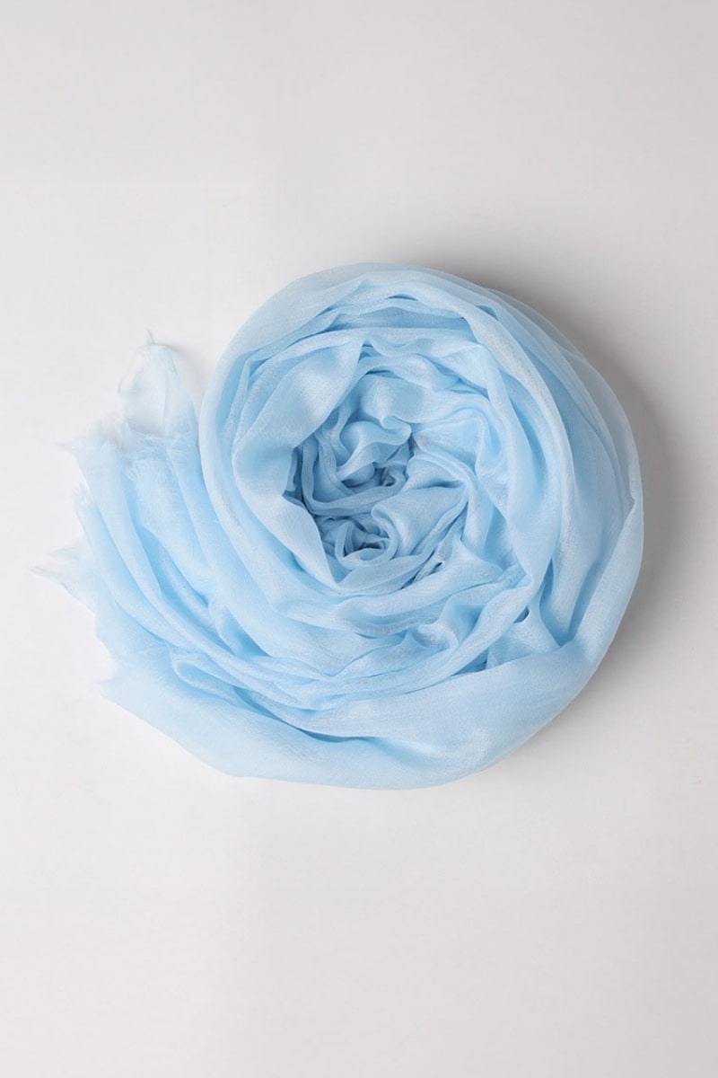 The looselyboho Sky Mist 100% Cashmere Scarf, in sheer light blue with softly frayed edges, is rolled into a spiral on a plain white background, highlighting its lightweight and delicate material.