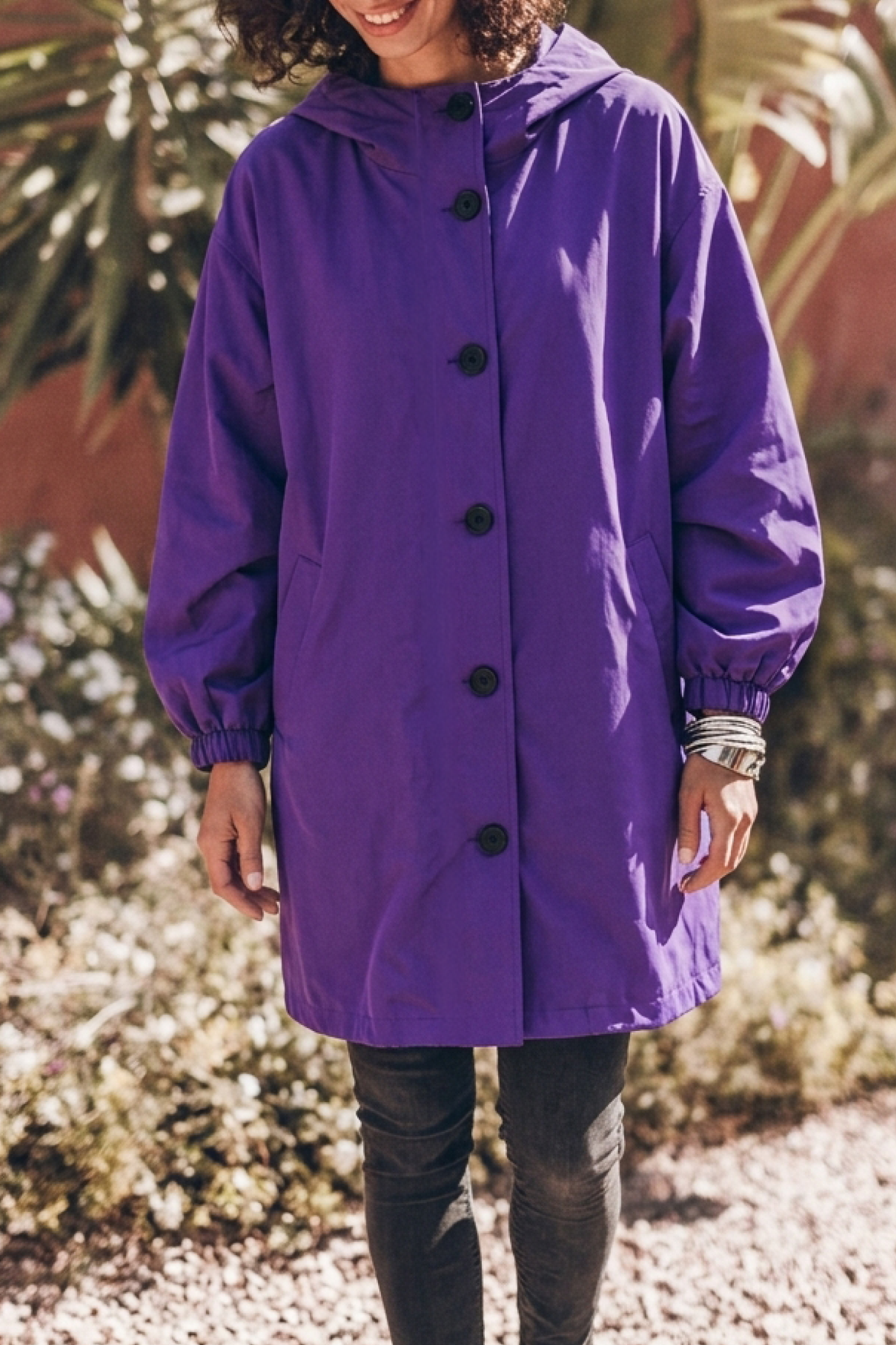 A person stands outdoors wearing the Icon Rain Windbreaker Jacket—a loose-fitting, long-sleeved purple rain jacket with black buttons—paired with dark pants and bracelets. The background features plants and gravel, and the person’s face is partially out of frame.