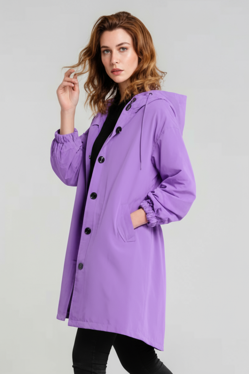 A person with wavy, shoulder-length hair stands against a light background wearing the looselyboho SOFT LAVENDER WINDBREAKER, which features an adjustable hood and black buttons, paired with black pants.