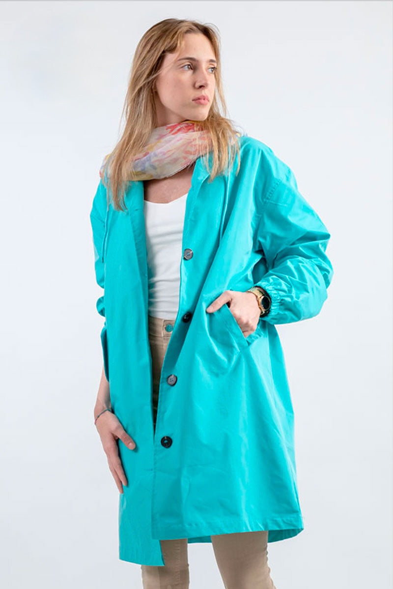 A person with long, light hair wears the looselyboho ELECTRIC CYAN WINDBREAKER with an adjustable hood over a white top, beige pants, and a colorful scarf, standing with one hand in the pocket against a plain background.