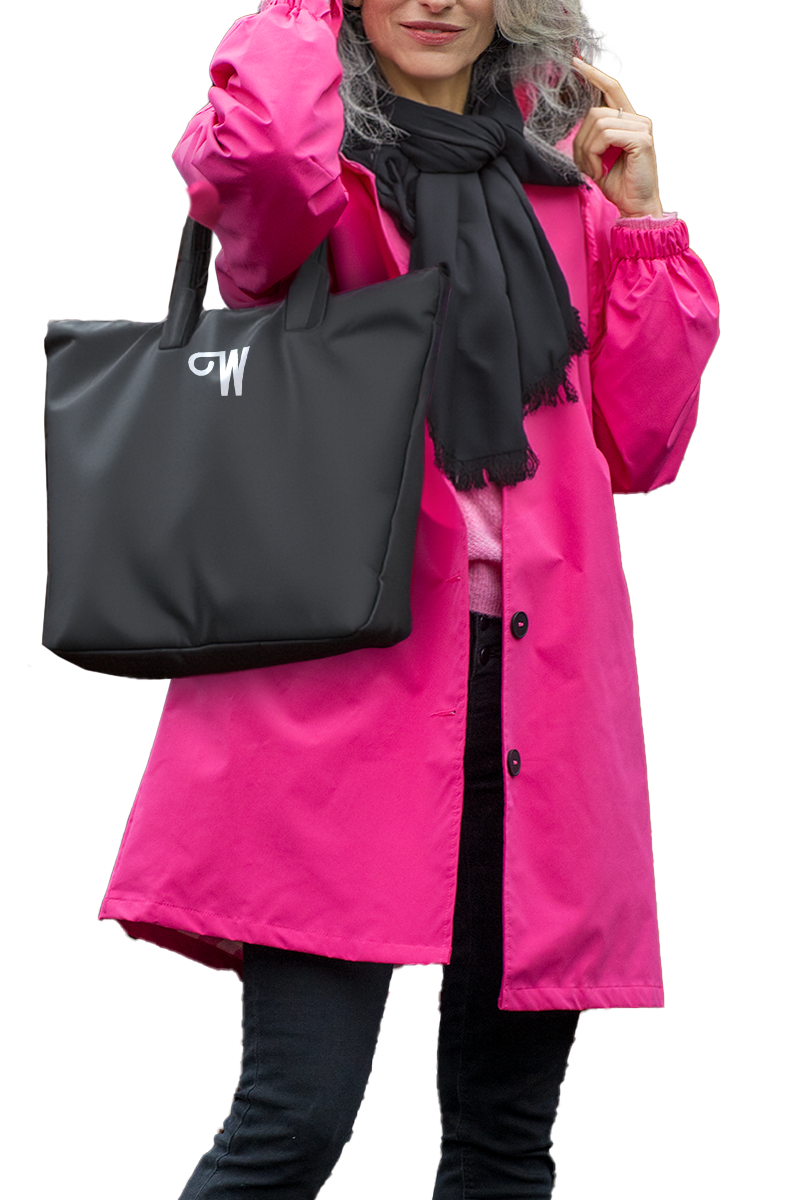A person is wearing an iconic looselyboho BARBARA PINK WINDBREAKER OUTFIT, black scarf, and black pants. They are holding the handle of a black tote bag with a white "GW" logo printed on it. The person is partially showing their face and has gray hair. The background is white.