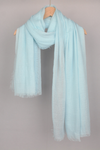 A light blue, semi-sheer all-weather "Color-POP Scarf" by looselyboho hangs on a wooden hanger against a plain gray background. The scarf has a slightly crinkled texture with frayed edges, draped in a simple, casual manner.