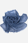 A lightweight crinkled blue Lightweight Linen Scarf from looselyboho is artfully arranged in a spiral formation, resembling a rose. The scarf has frayed edges, giving it an artisanal charm and textured appearance. The plain white background emphasizes the scarf's color and texture.