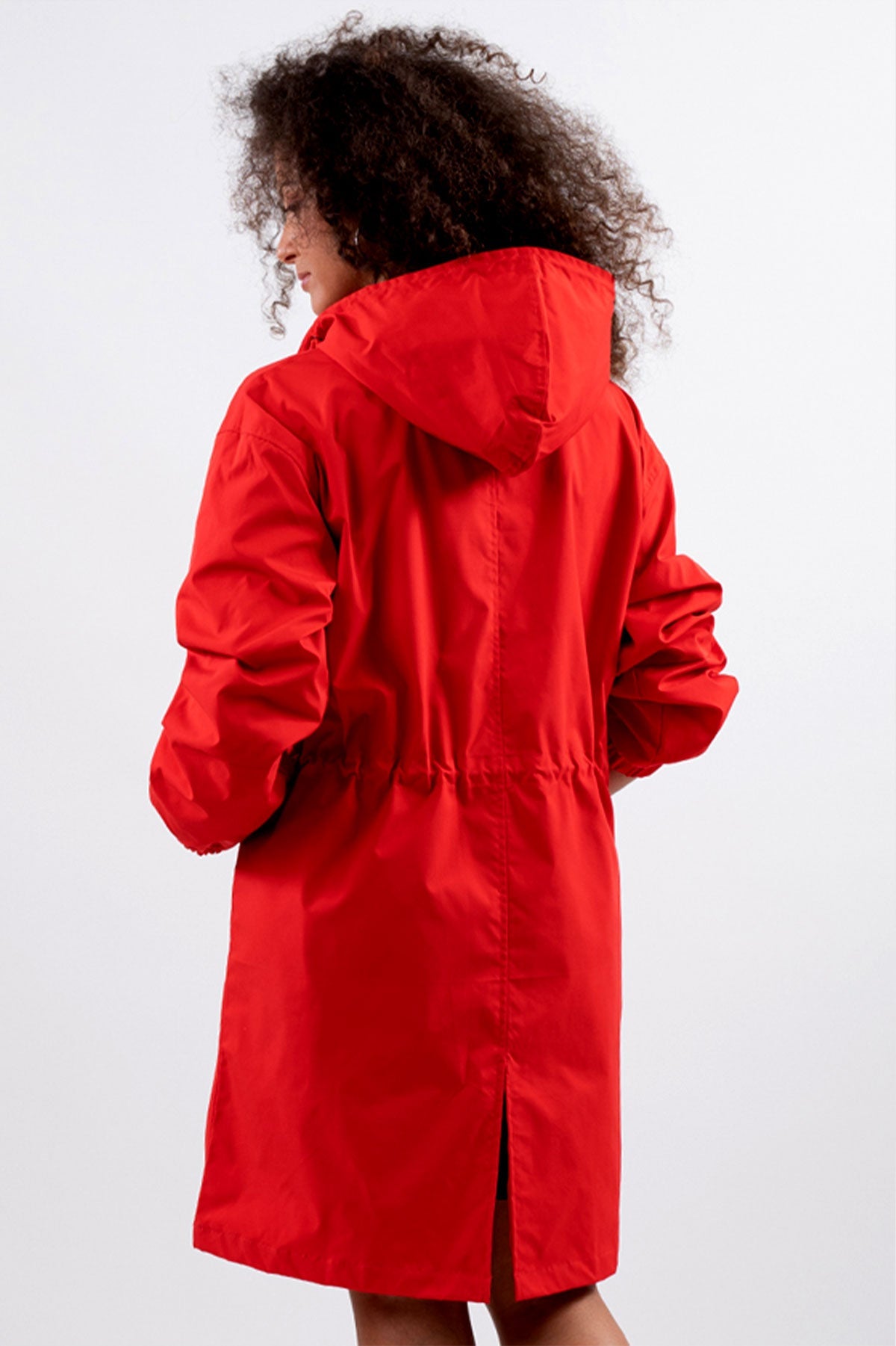 Person with curly hair shown from the back, wearing a bright red Water Resistant Oversized Hooded Windbreaker Rain Jacket by The WJacket. The jacket, featuring an adjustable waistband and falling just above the knees, combines timeless style and versatility. The background is plain and light-colored.