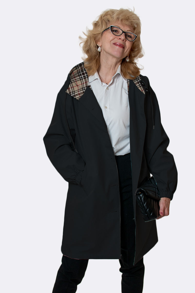 A blonde woman in glasses wears the looselyboho LUXE BLACK WATERPROOF WINDBREAKER with a plaid-lined hood, paired with a white shirt and dark pants. She holds a black clutch and smiles against a plain light background.