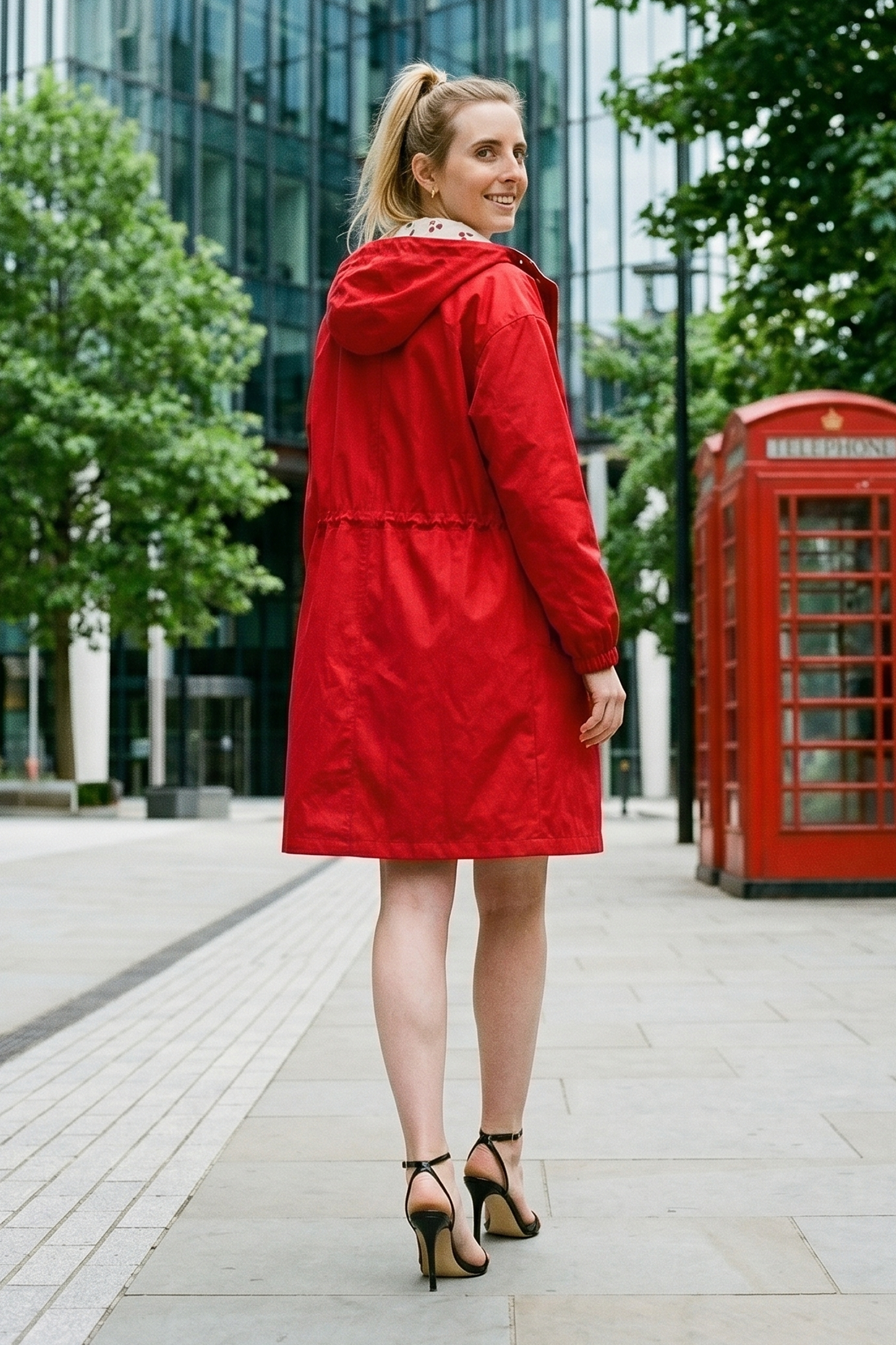 Wearing The Water Resistant Hooded Oversized Windbreaker Rain Jacket and black heels, a person with a ponytail stands on a city sidewalk, glancing over their shoulder. Behind them are trees, a glass building, and a red phone booth.