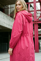 A person with long blonde hair stands outdoors in front of a modern building, wearing the Icon Windbreaker Rain Jacket in bright pink and looking over their shoulder at the camera.