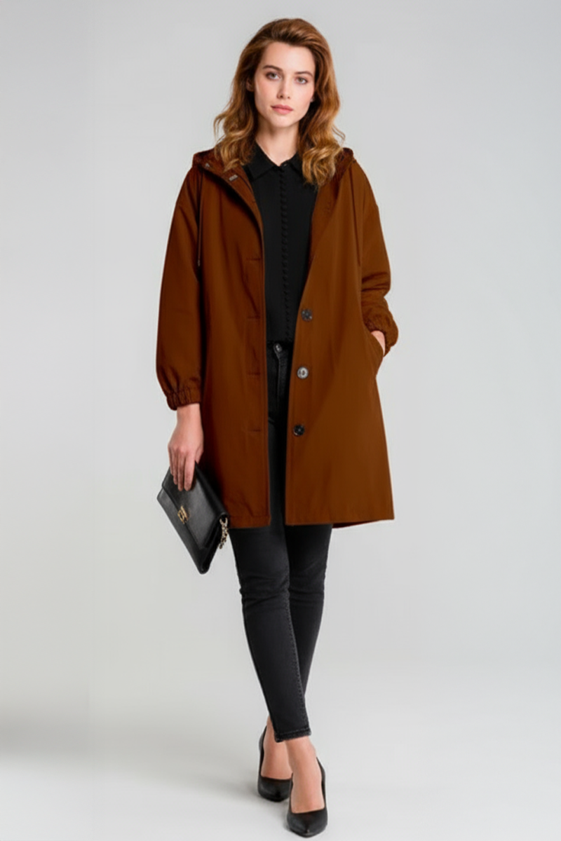 A woman stands against a plain background wearing the looselyboho BURNT BROWN WINDBREAKER with an adjustable hood over a black shirt and pants, holding a black clutch, and completes the look with sleek black heels.