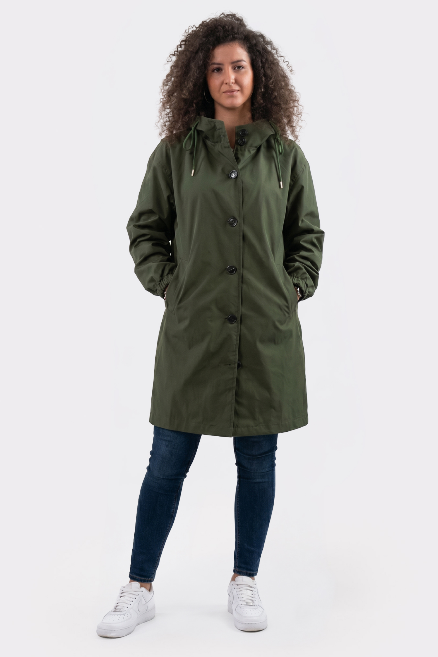 A person with curly hair stands facing forward against a plain white background, wearing The Water Resistant Hooded Oversized Windbreaker Rain Jacket in olive green, paired with dark blue jeans and white sneakers, hands in jacket pockets.