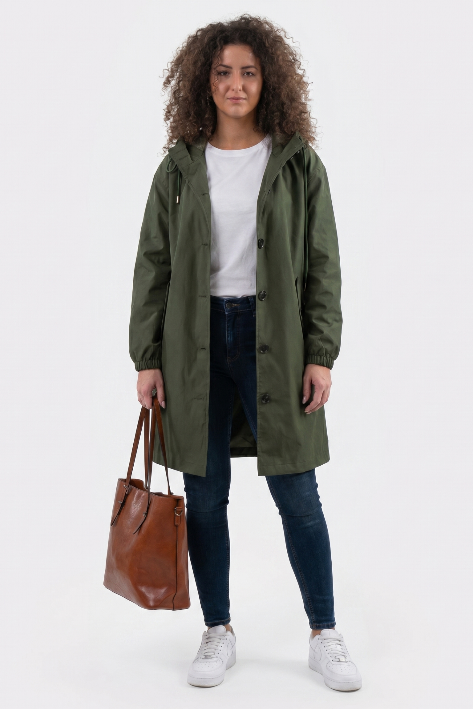 A person with curly hair wears The Water Resistant Hooded Oversized Windbreaker Rain Jacket in green, a white t-shirt, dark jeans, and white sneakers, holding a brown leather tote bag and facing the camera against a plain white background.