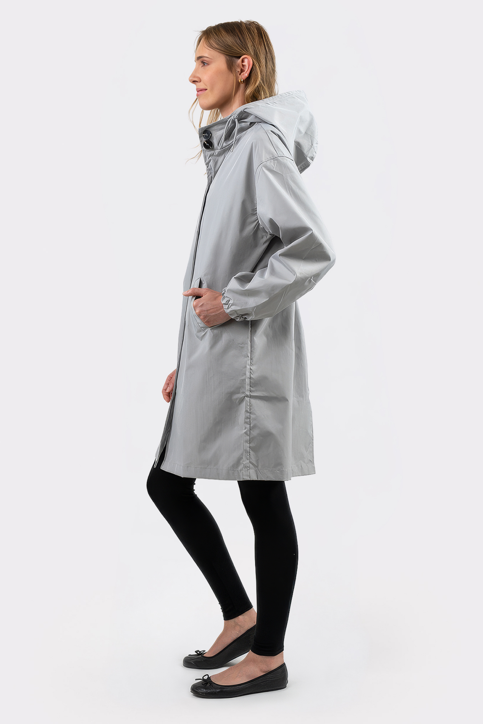 A person stands in profile wearing The Water Resistant Hooded Oversized Windbreaker Rain Jacket in light gray, paired with black leggings and ballet flats, against a plain white background.