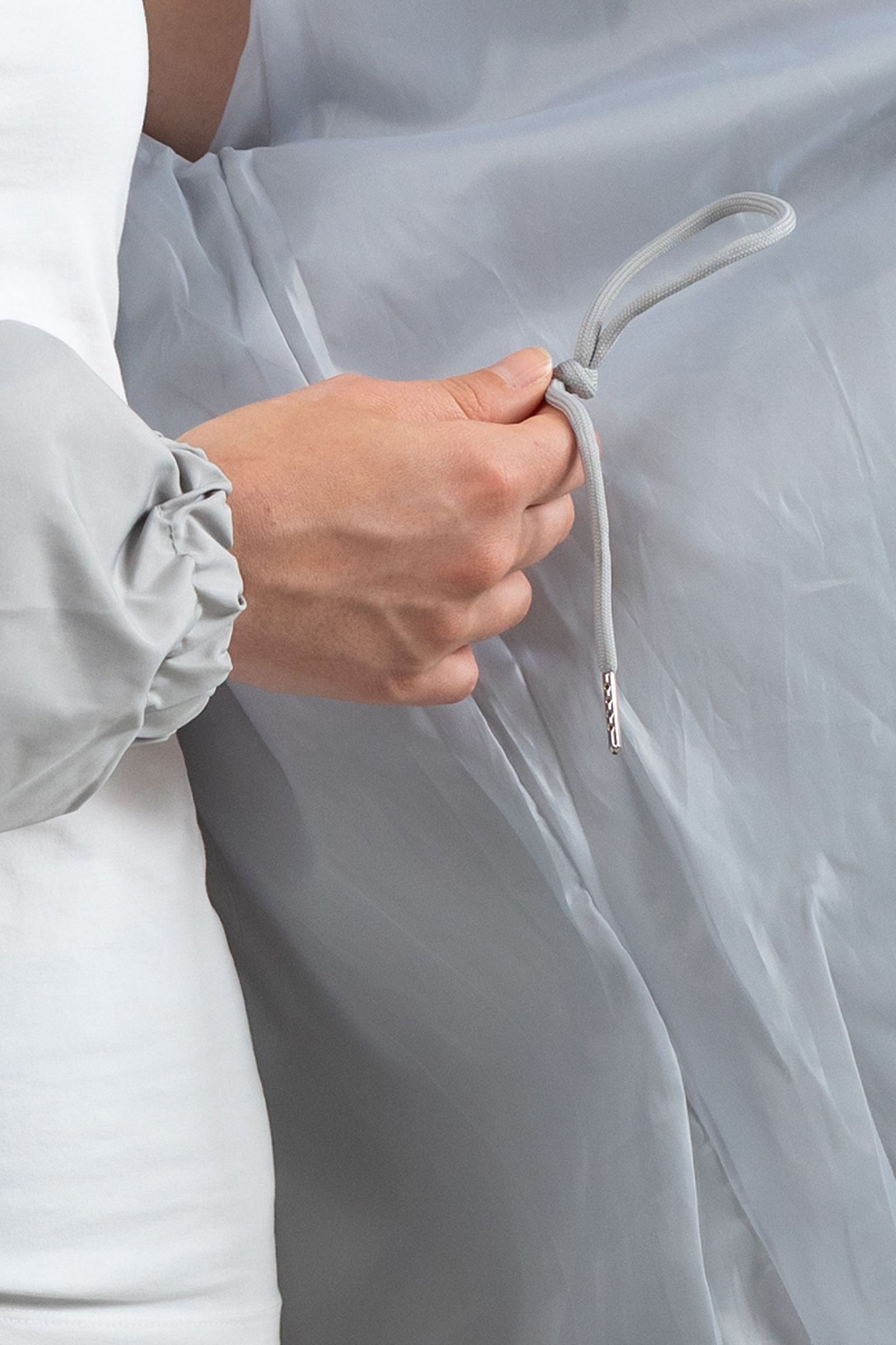A person in The Water Resistant Hooded Oversized Windbreaker Rain Jacket, featuring a light gray color and ruched sleeves, holds a knotted gray drawstring against a semi-transparent light gray fabric background.