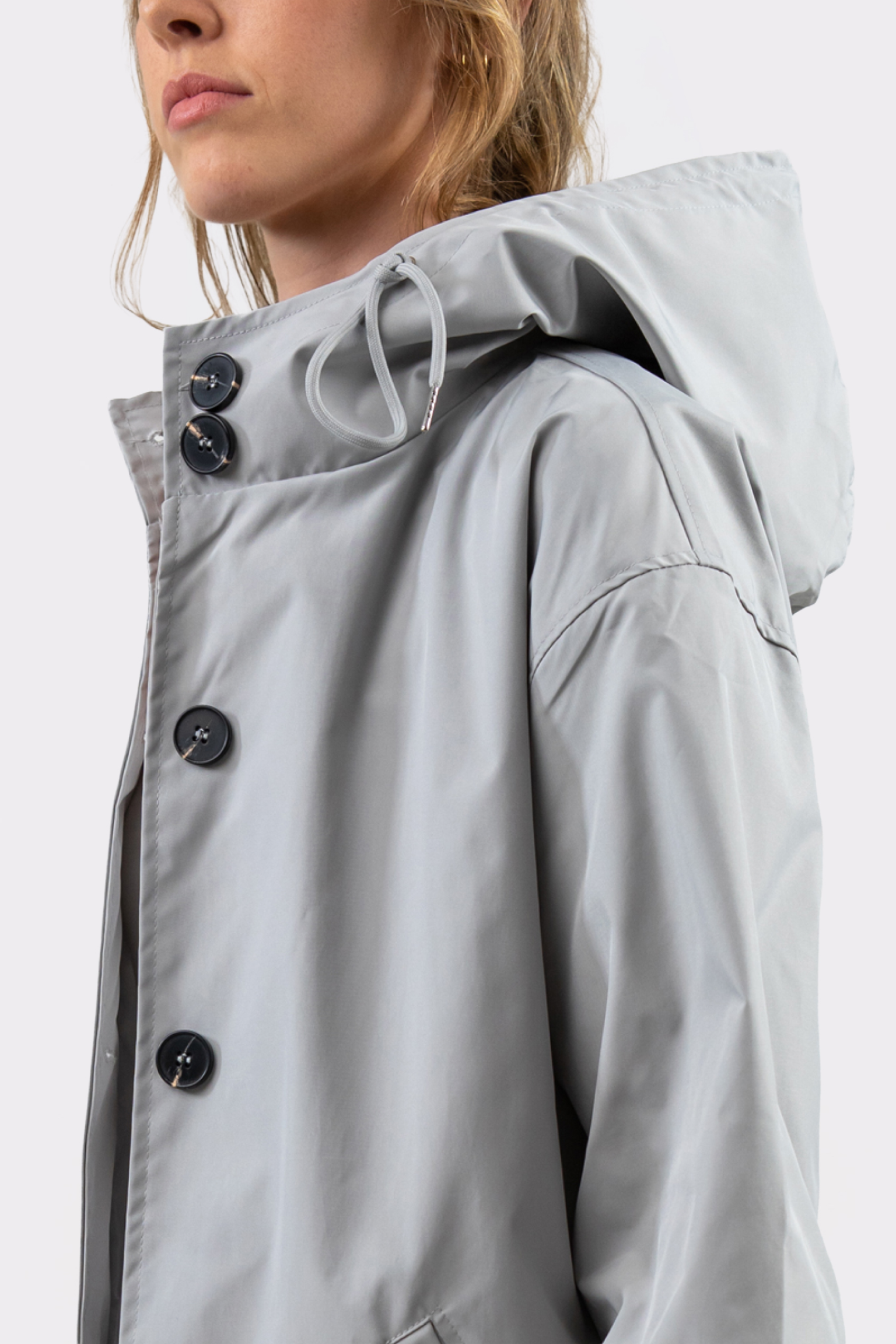 A person wearing The Water Resistant Hooded Oversized Windbreaker Rain Jacket in light gray, partially unbuttoned, with large black buttons. The person’s face is partly visible from the shoulders up against a plain background.