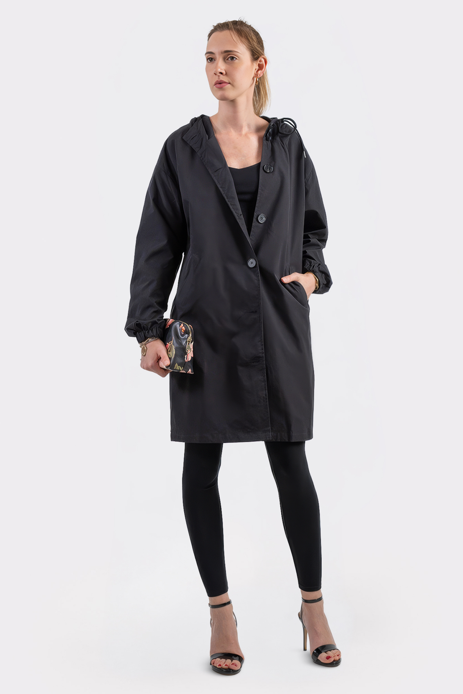 A person stands facing forward, wearing the Icon Windbreaker Rain Jacket over black leggings and top, paired with a graphic clutch and black high-heeled sandals, set against a plain white background.