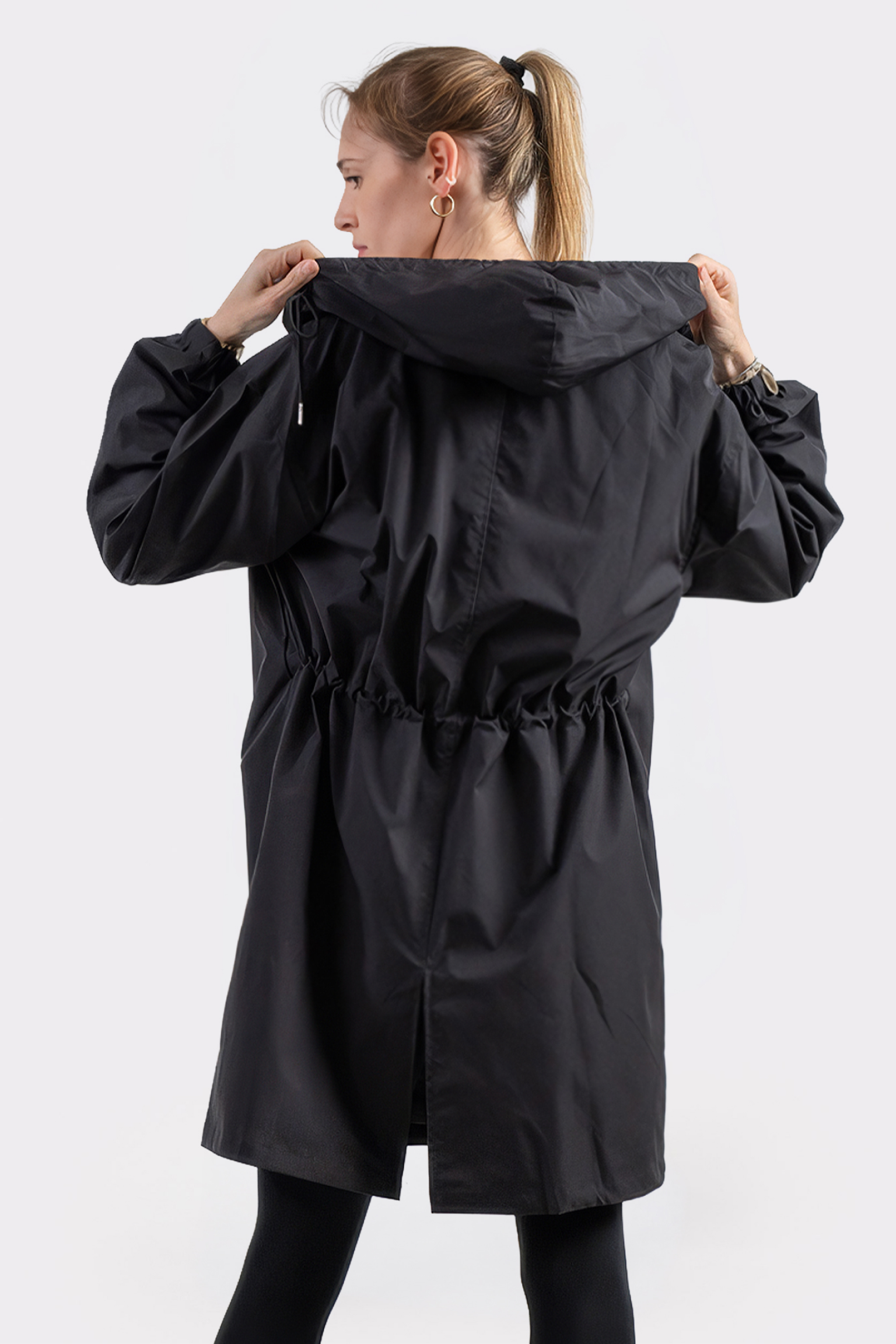 A woman with light hair in a ponytail is seen from behind, wearing The Water Resistant Hooded Oversized Windbreaker Rain Jacket in black. She holds the hood with both hands against a plain white background.