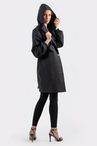 A person wearing the Icon Windbreaker Rain Jacket, black leggings, and high-heeled sandals stands against a plain white background, looking at the camera while holding the edge of the jacket’s hood with one hand.