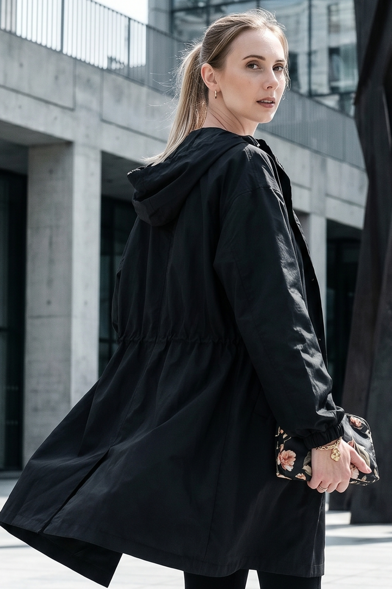 A person with light hair in a ponytail is wearing the Icon Windbreaker Rain Jacket and holding a floral clutch while standing outdoors in front of a modern concrete building.