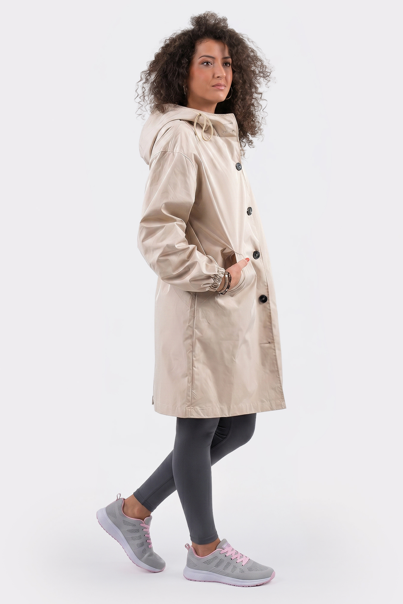 A person stands sideways against a plain white background wearing The Water Resistant Hooded Oversized Windbreaker Rain Jacket, paired with dark gray leggings and gray athletic shoes with light pink accents.