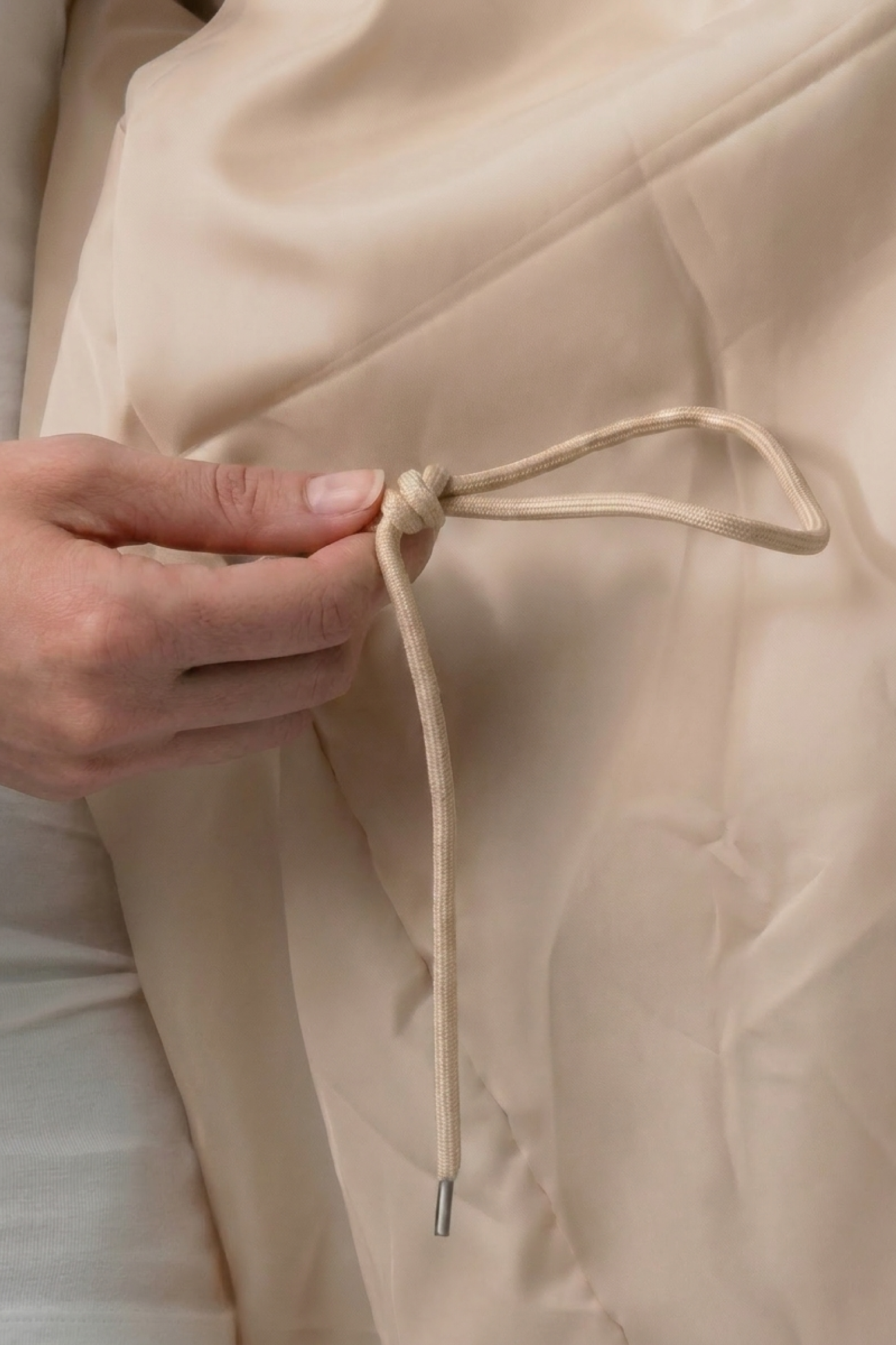 A hand is tying a beige string into a bow on The Water Resistant Hooded Oversized Windbreaker Rain Jacket, adjusting the fit of the light-colored outerwear.