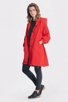 A woman with light brown hair and a neutral expression faces forward, wearing the Icon Windbreaker Rain Jacket in bright red, paired with a black button-up shirt, black skinny jeans, and black slip-on shoes featuring a tan heel.