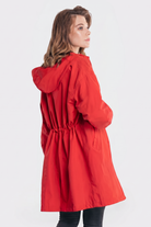A person in the Icon Windbreaker Rain Jacket, featuring a bright red color, hood, elastic waist, and two side pockets, stands at a slight angle facing away and looks over their shoulder against a plain white background.