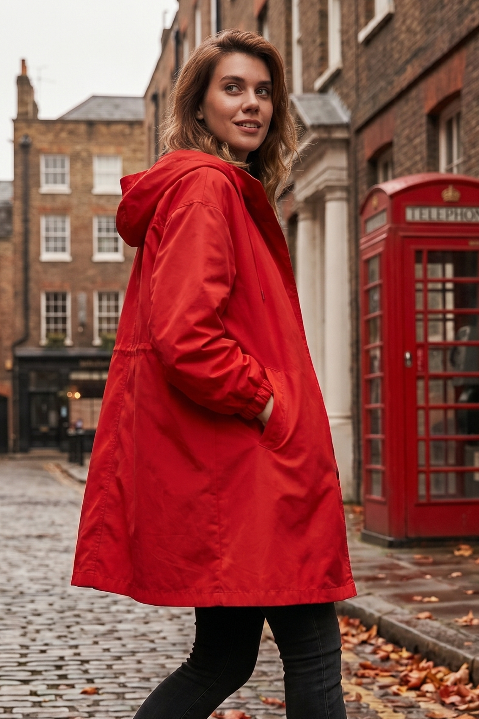 A woman wearing the Icon Windbreaker Rain Jacket stands on a cobblestone street beside a red British phone booth, with brick buildings and autumn leaves in the background.