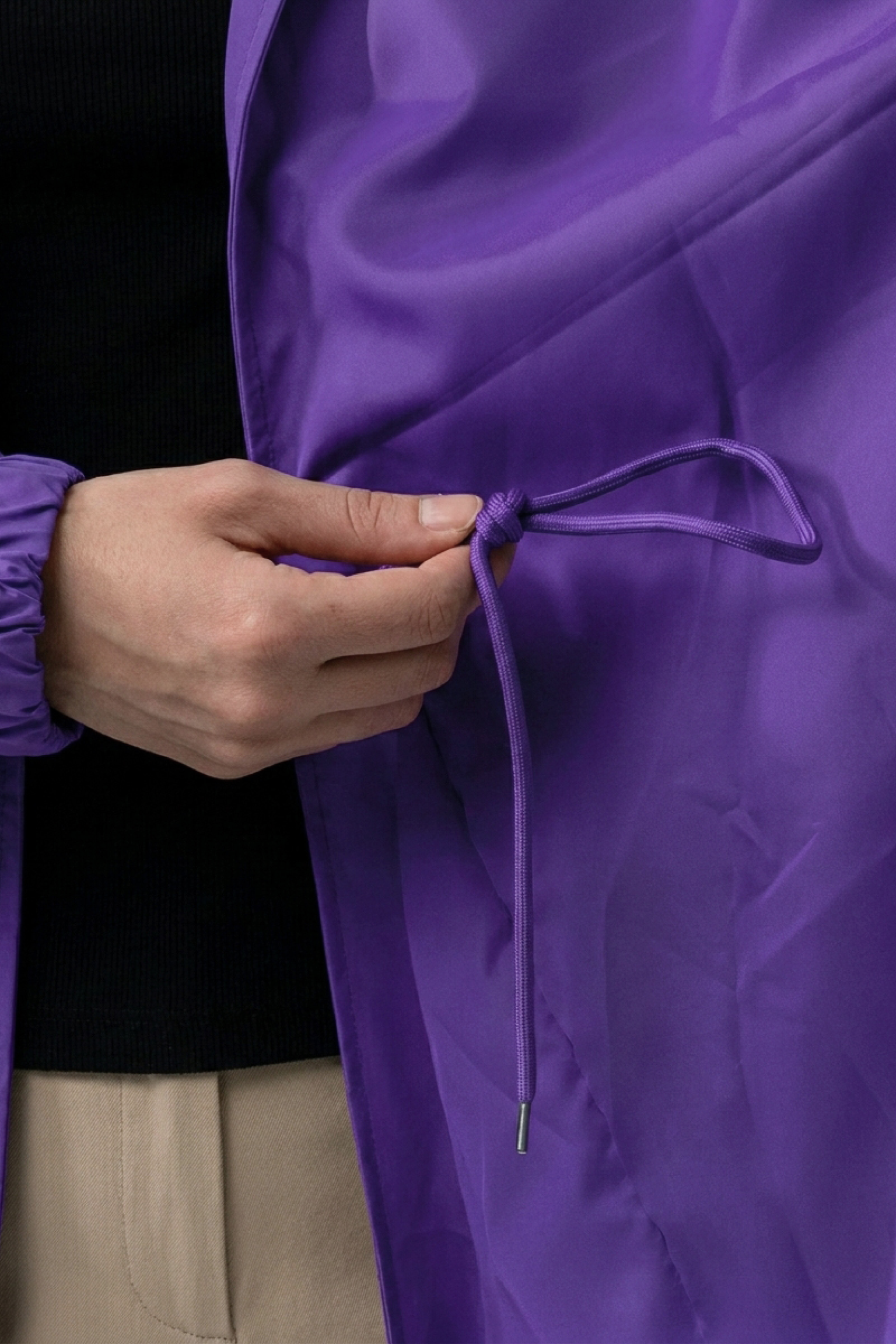 A person adjusts the drawstring on an Icon Windbreaker Rain Jacket in purple, focusing on the hand holding the string. Part of the jacket sleeve and beige pants are also visible.