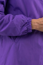 A person is shown from shoulder to waist wearing the Icon Windbreaker Rain Jacket in purple, featuring elastic cuffs, a side pocket seam, and one hand partially visible.