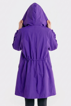 A person with their back to the camera wears the Icon Windbreaker Rain Jacket in purple, hood up and hands holding the sides. The background is plain and light-colored.