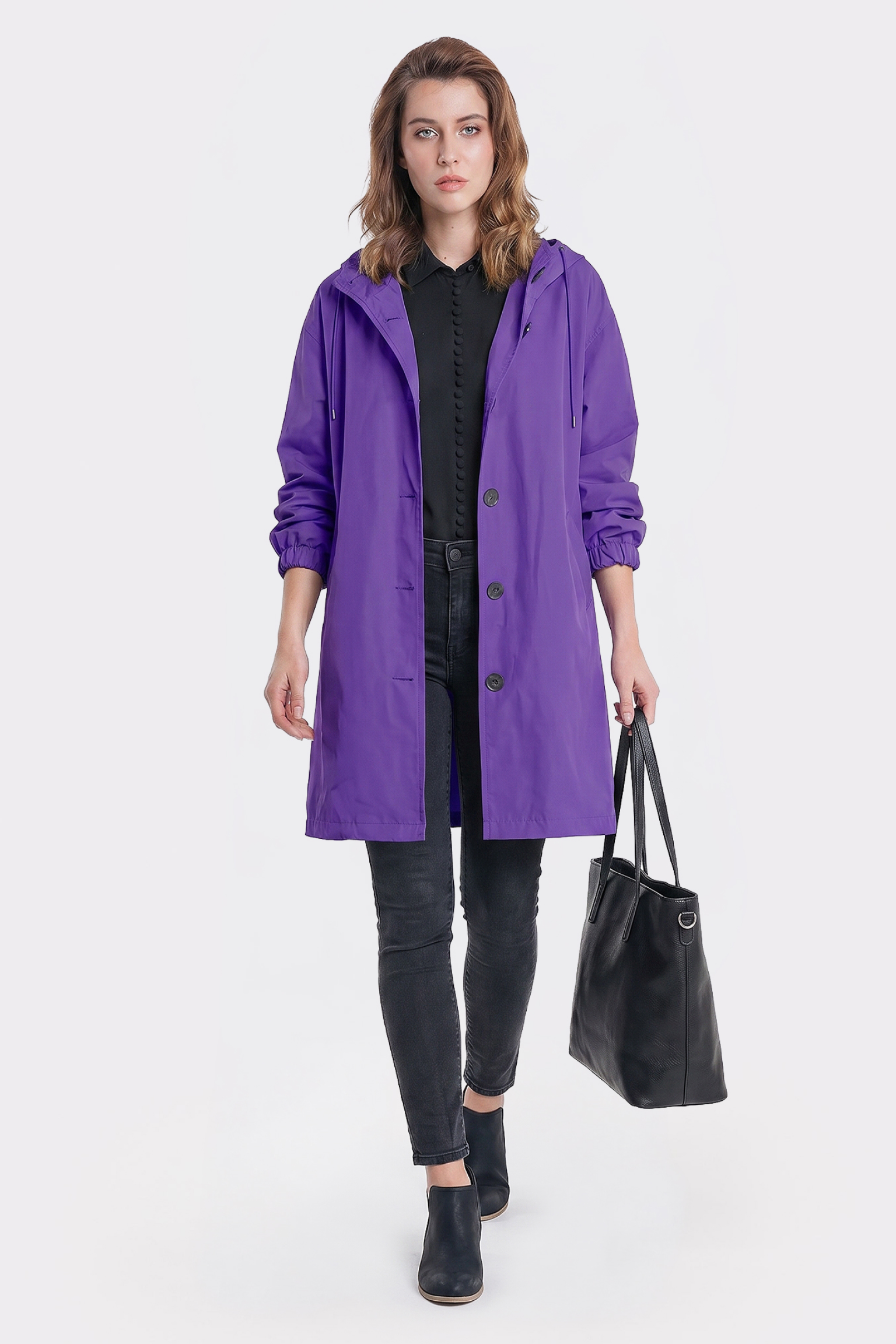 A person wearing the Icon Windbreaker Rain Jacket in purple over a black shirt and pants, holding a black tote bag, stands against a plain white background.