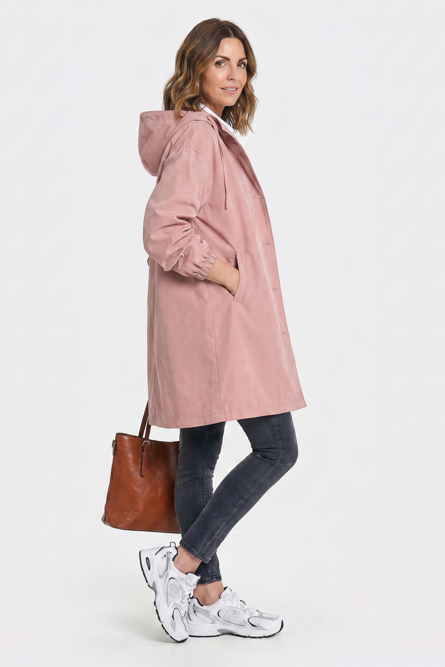 A woman in profile wears the Icon Windbreaker Rain Jacket in pink, paired with dark skinny jeans and white sneakers. She holds a brown leather handbag, smiling slightly at the camera against a plain white background.