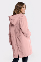 A woman with wavy brown hair is seen from the side and back, wearing the Icon Windbreaker Rain Jacket in light pink, mid-thigh length, standing against a plain light background with her hands relaxed by her sides.