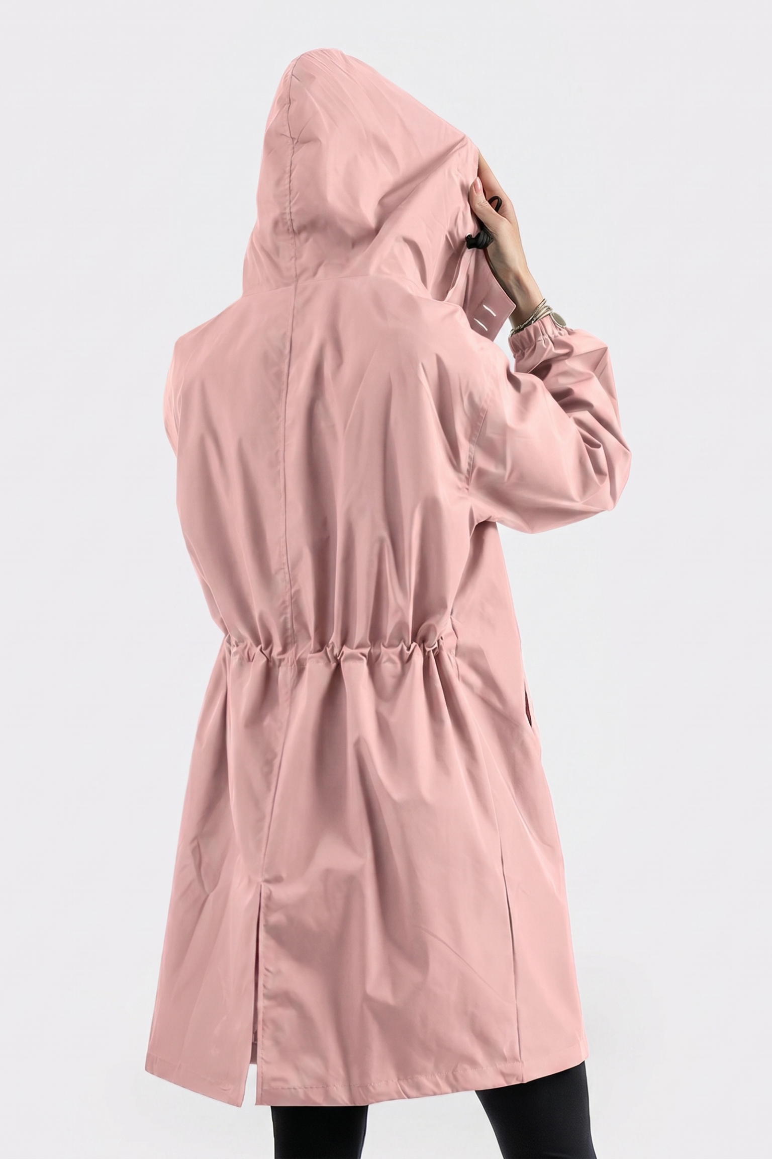 A person stands facing away from the camera, holding the hood of The Water Resistant Hooded Oversized Windbreaker Rain Jacket, a light pink raincoat with an elastic waistband. The background is plain white.