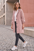 A woman smiles while walking on a cobblestone street, wearing the Icon Windbreaker Rain Jacket with dark jeans and white sneakers. One hand is in her pocket, and modern concrete buildings appear in the background.