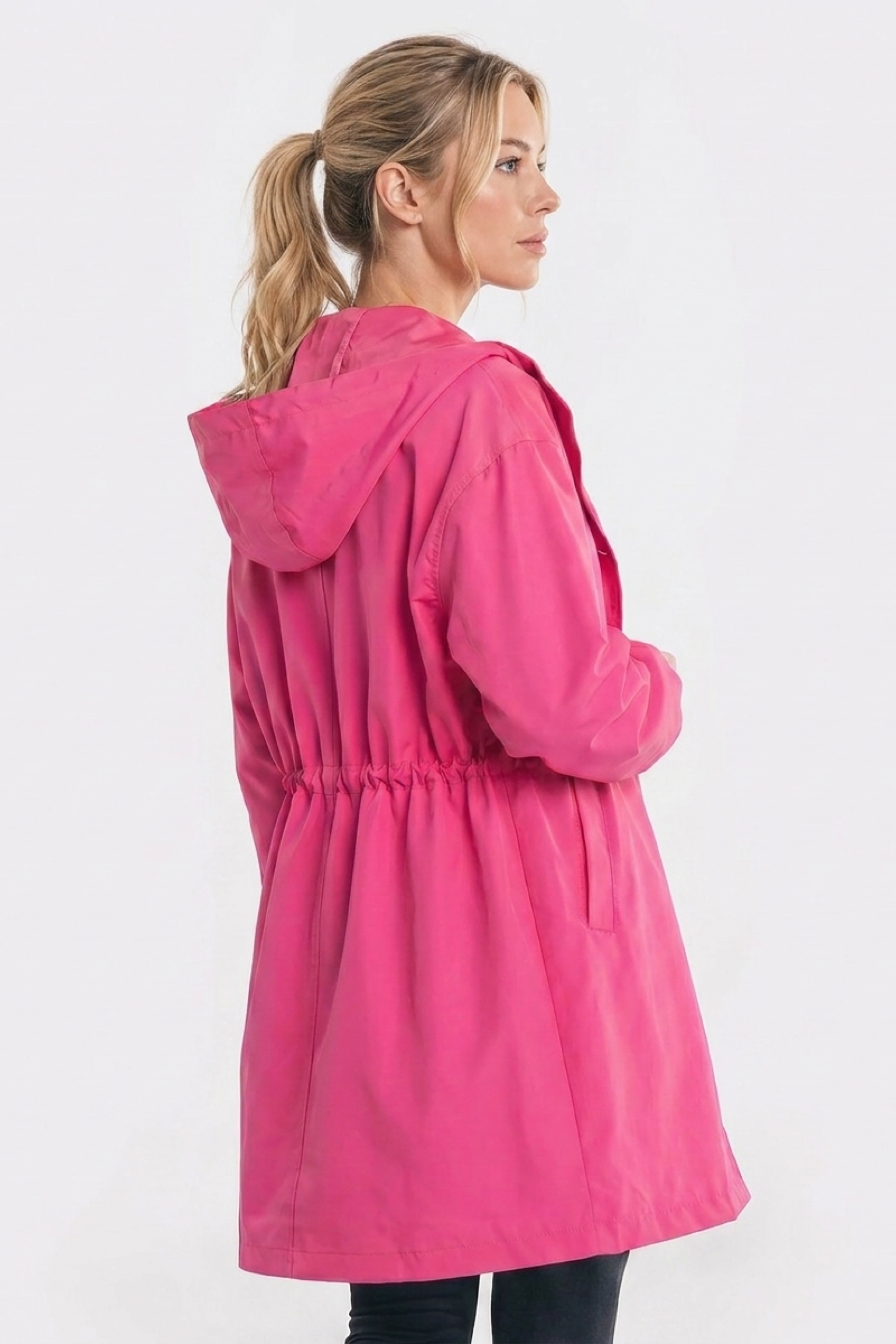 A person with blonde hair in a ponytail wears The Water Resistant Hooded Oversized Windbreaker Rain Jacket in bright pink, featuring an elastic waist, standing sideways facing left against a plain white background.