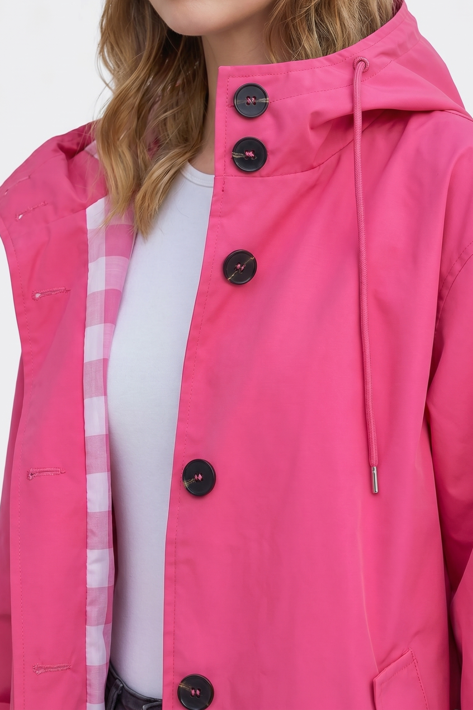 A person wears The Water Resistant Hooded Oversized Windbreaker Rain Jacket in bright pink with black buttons and a hood over a white shirt. The coat’s inner lining features a pink and white checkered pattern. Only the lower face and upper torso are visible.