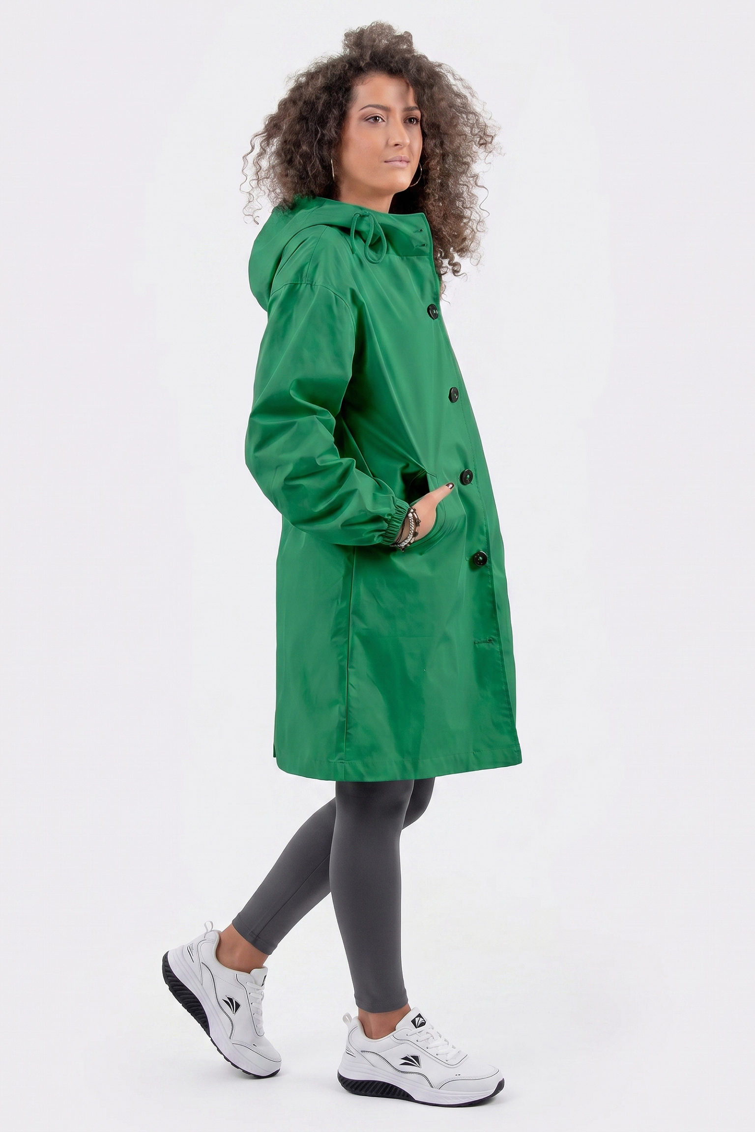 A person with curly hair wears The Water Resistant Hooded Oversized Windbreaker Rain Jacket in green, paired with gray leggings and white sneakers. They stand sideways with one foot slightly raised against a plain white background.