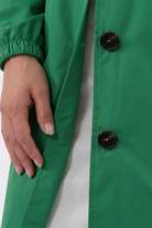 A close-up of a hand resting on the side of the green Icon Windbreaker Rain Jacket with large black buttons, worn over a white ribbed garment.