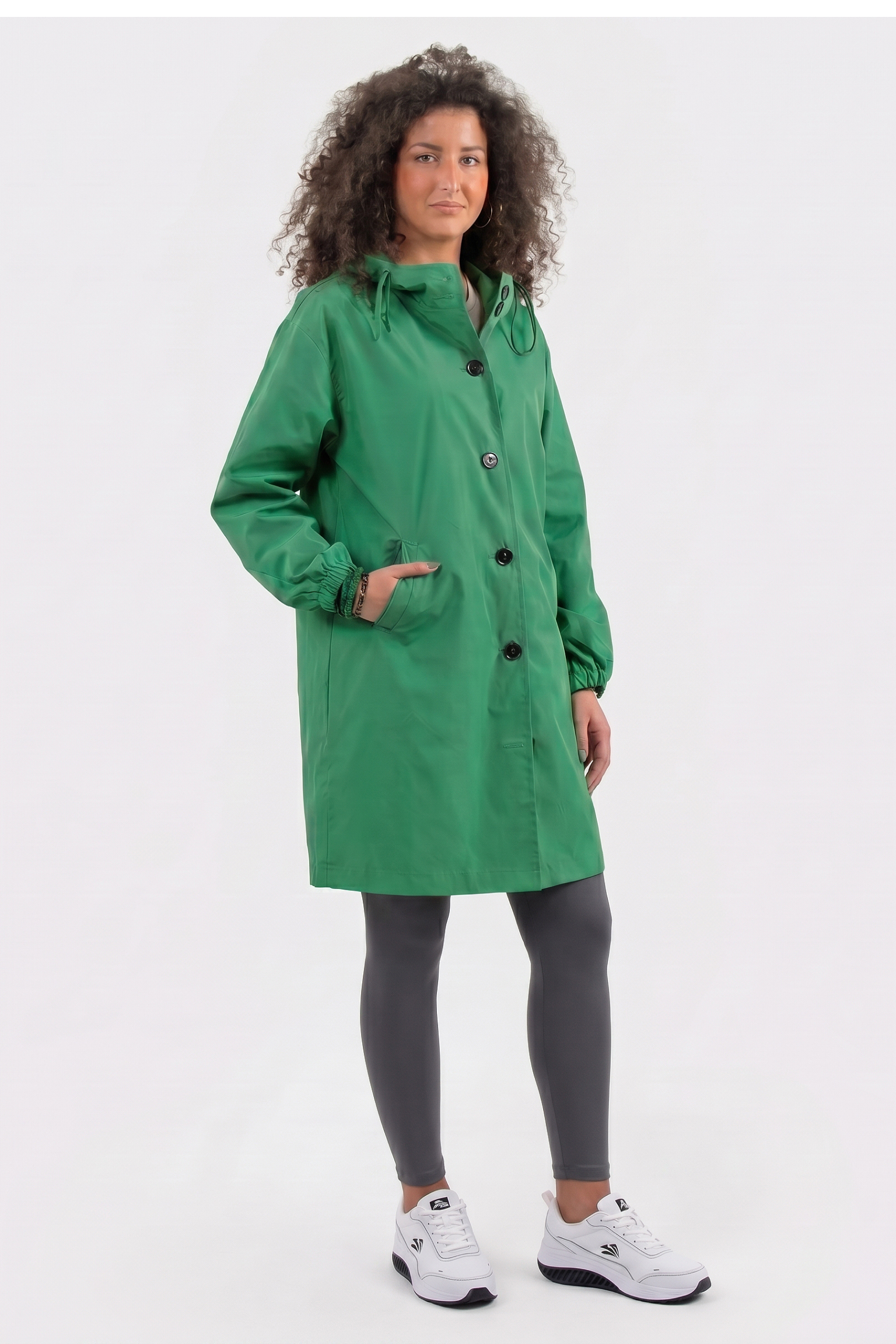 A person with curly hair wears the Icon Windbreaker Rain Jacket in green, paired with gray leggings and white sneakers, standing against a plain light background with one hand in their pocket.