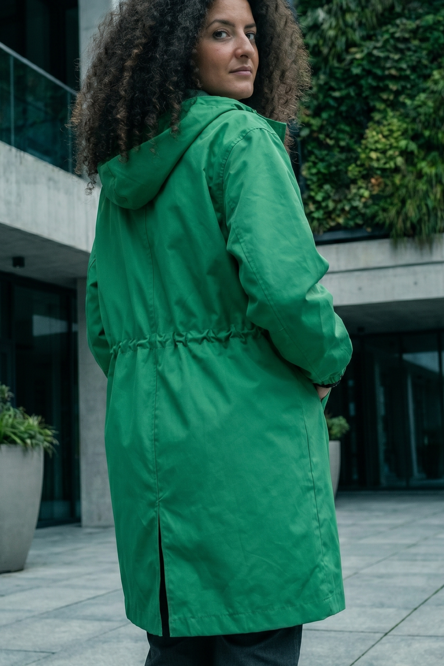 A person with curly hair wears The Water Resistant Hooded Oversized Windbreaker Rain Jacket in green, standing on a paved area by a modern glass-front building with plants. They face away while looking over their shoulder.