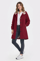 A person stands against a plain background, smiling with one hand in the pocket of the red Icon Windbreaker Rain Jacket, worn over a white button-up shirt, dark gray jeans, and white sneakers.