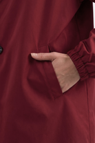 A person wearing the Icon Windbreaker Rain Jacket, which has a maroon color and elastic cuffs, has their right hand in the jacket’s side pocket.