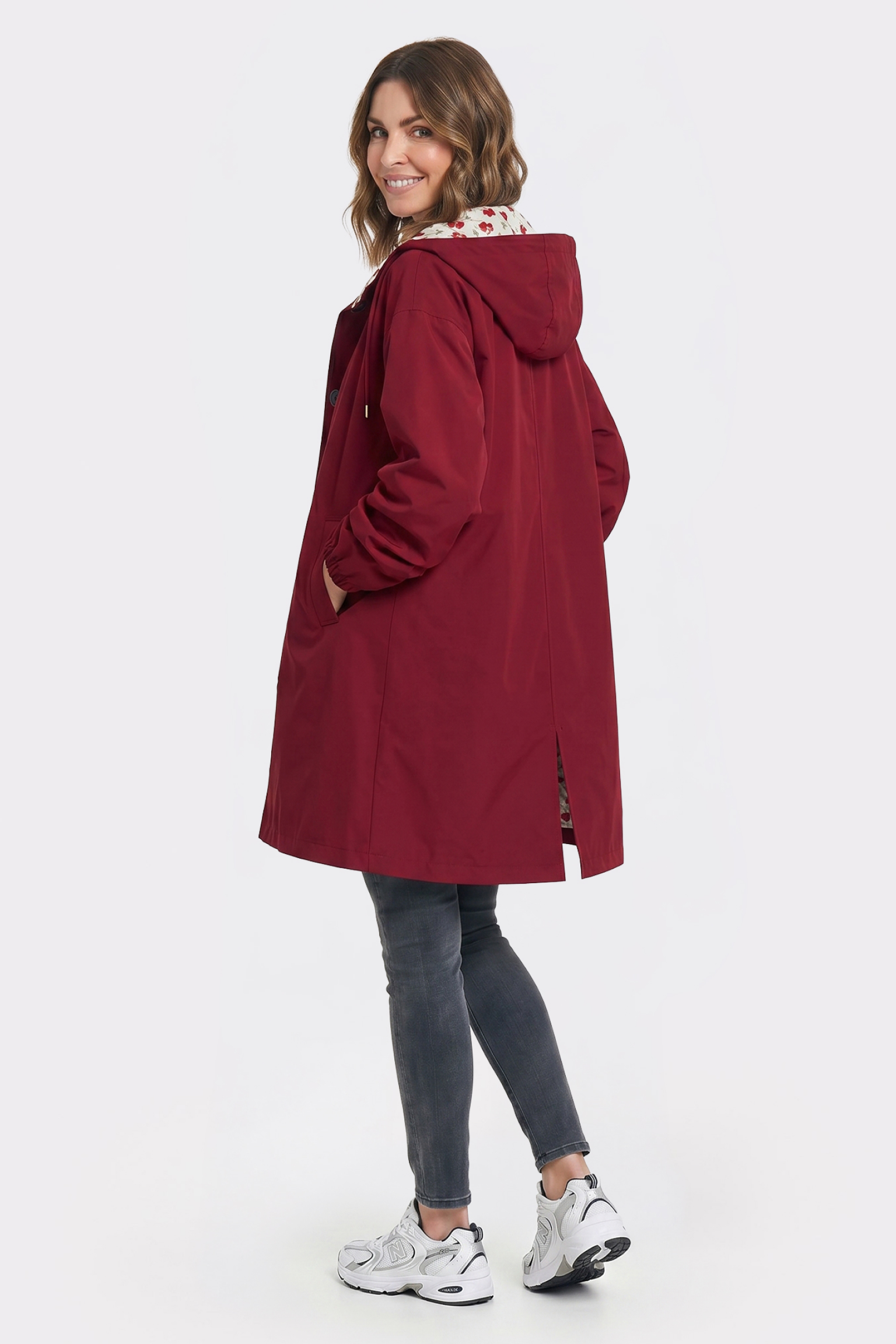A woman stands facing away and looks back at the camera, wearing the Icon Windbreaker Rain Jacket in red, paired with dark gray jeans and white sneakers against a plain white background.