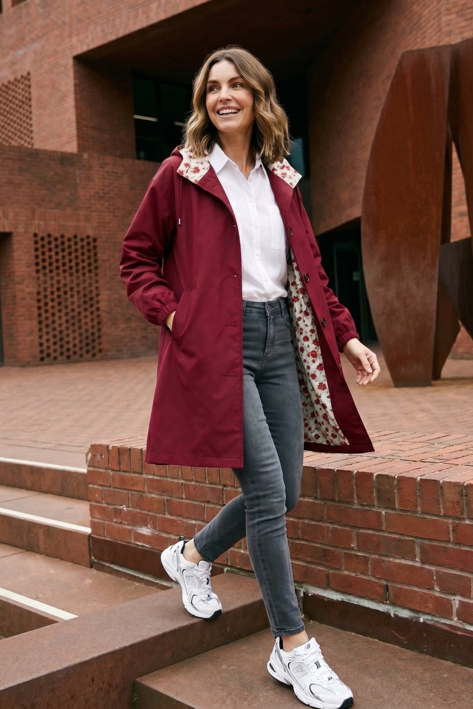 A woman wearing The Water Resistant Hooded Oversized Windbreaker Rain Jacket, a white shirt, gray jeans, and white sneakers walks outdoors on steps by a brick building with her hands in her jacket pockets, smiling.